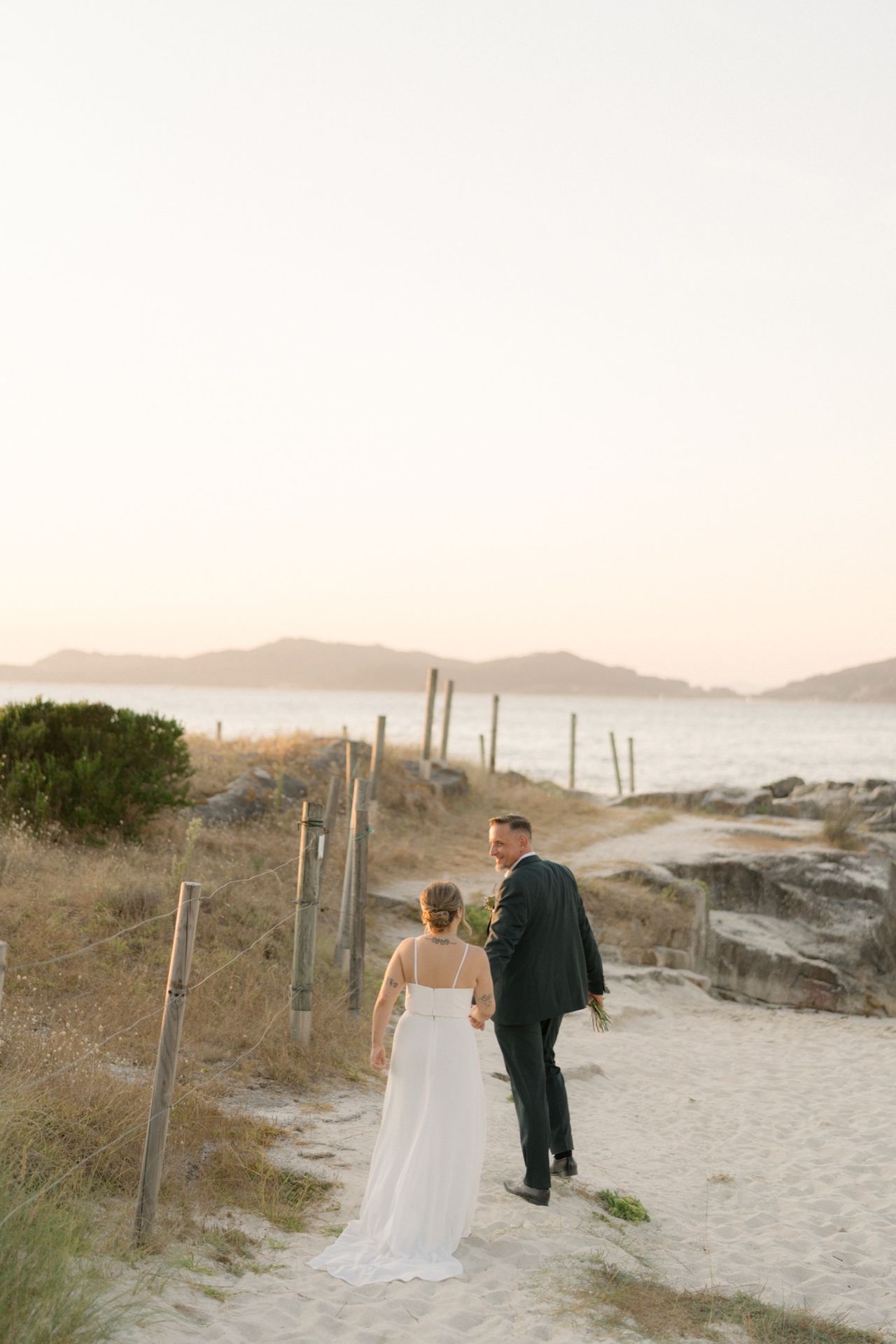 Pareja de novios paseando de la mano por el camino hacia la playa al atardecer, con la ría de Vigo al fondo